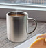 A stainless steel coffee mug with steam rising from the hot beverage inside, positioned near a plate of pancakes.