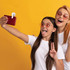 A selfie is being taken by two girls wearing sunglasses and white t-shirts, with a bright yellow background.