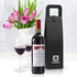 A black wine carrier beside a bottle of red wine and two glasses filled with wine, with tulips in the background.