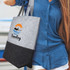 A grey and black tote bag with a logo, held by a person wearing a blue shirt, featuring a sunset and waves design.