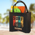 A black jute tote bag with a logo, filled with a towel and green flip-flops on a sandy background.