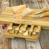 A wooden cheese board with cheese slices, scattered pieces, and four cheese knives, all set on a rustic wooden surface.