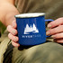 A blue enamel mug held by a person, featuring a logo with trees and a winding path.