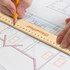 A wooden 30cm ruler rests on a blueprint with a hand holding a pencil. The ruler features a logo.
