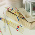 A wooden box containing multicoloured pick up sticks, scattered on a light surface. The box has a logo.