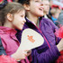 A foldable event fan in cream, held by a girl, with a logo displayed, surrounded by cheering spectators.