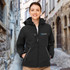 A woman wearing a black softshell jacket with a hood, featuring a logo on the chest, stands in a cobblestone alley.