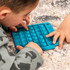 A turquoise sensory board with multiple bubble-like buttons, being used by two children on a textured surface.