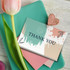 A thank you card with a teal and peach abstract design, resting on a green surface with a pink envelope and a tulip.
