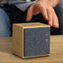 A square, bamboo Bluetooth speaker features a grey fabric front and volume controls, sitting on a wooden table.