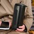 A black Bluetooth speaker with a textured surface, held by a person alongside a smartphone.