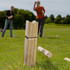 A wooden Kubb game set on grass, featuring light-coloured wooden blocks and a player in the background.