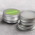 A silver tin for lip balm with a green decorative lid, featuring a logo. Two tins are partially open on a grey fabric background.