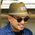 A straw Santiago Fedora hat in natural tones, featuring a logo patch, worn by a smiling person with sunglasses.