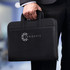 A black travel portfolio case with two handles, featuring a logo on the front, held by a person in a suit.