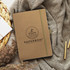 A brown notebook with a logo, lying on a textured fabric beside pens and decorative elements.