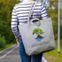 A grey tote bag featuring a tree design and a logo, carried by a person in a striped shirt on a pathway.