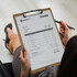 A wooden clipboard holds an invoice with a logo at the top, held by a person writing with a pen.