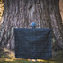 A black puffer blanket is held up by a person in front of a large tree. The scene is outdoors.