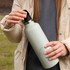A drink bottle in a soft green colour with a carry lid, held by a person wearing a light jacket in an outdoor setting.