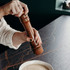 A wooden pepper mill being operated by a person, with a bowl of creamy mixture on a dark surface.
