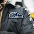 A grey jacket with a custom woven patch featuring a vehicle and mountains in blue and white.
