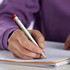 A green recycled plastic pen with a red cap, held by a hand, writing in a notepad. The pen features a logo.