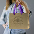 A medium brown paper bag with purple ribbon handles, featuring a logo on the front.