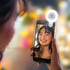 A selfie light is attached to a smartphone, illuminating a smiling person with a blurred background of lights.