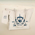 A canvas tote bag in cream with a nautical design and rope handles, featuring a matching tag.
