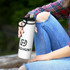 A drink bottle in white with a black lid and handle, featuring a logo. The bottle is held by a person in casual attire.
