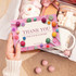 A5 message card in pink, decorated with colourful candies, held in a hand against a background of sweets and gifts.