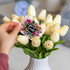 A hand holds a pink A7 greeting card featuring floral designs, placed among white tulips in a vase.