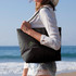 A black tote bag is held by a woman in a beach setting, featuring a logo on the front.