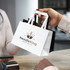 A small flat handle paper bag in white with a logo, held by a person at a counter.