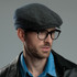 A flat cap in a herringbone pattern, predominantly grey, worn by a model with glasses. Features a logo.
