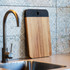 A wooden serving board with a black handled edge, leaning against a tiled wall near a sink.