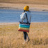 A person wearing a yellow beanie carries a multi-coloured striped picnic blanket against a natural landscape.