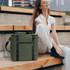 A soft-touch cooler bag in olive green sits beside a smiling woman on wooden steps. The bag features a zip and straps.