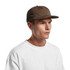 A brown cap with a curved brim, worn by a model in a white t-shirt. The cap features stitching details and a logo.