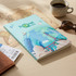 A teal notebook featuring travel-themed illustrations, placed on a wooden surface alongside a coffee cup and sunglasses.
