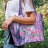 A cotton tote bag in shades of purple and pink, featuring a floral design and a logo.