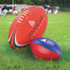 Two AFL balls on green grass; one red with a logo and one smaller red and blue with a logo.