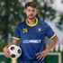 A man holds a soccer ball while wearing a blue and yellow custom men's soccer top with a logo.