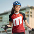 A woman wearing a red and black cycling jersey with a logo, holding a bicycle, and wearing a blue helmet.