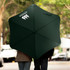A dark green sport umbrella with a logo, opened and being held by two people, against a blurred outdoor background.