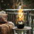A glass and metal burner with a flame, set on a wooden table outdoors beside a knitted blanket.