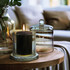 A glass candle in a clear holder with a lit wick and a cloche lid beside it on a wooden table.