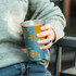 A drink cup in teal and yellow featuring colourful floral and citrus designs. It has a metallic interior and branding.