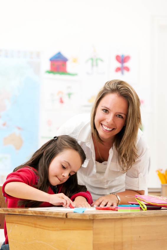 Image of female teacher smiling leaning over female student at desk on classroom background